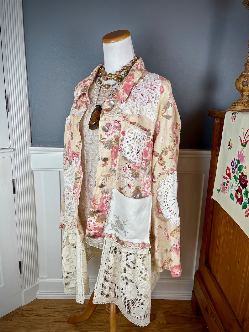 May include: A beige floral print jacket with pink and red flowers, white lace accents, and a white pocket. The jacket is displayed on a mannequin with a layered necklace and a wooden pendant. The jacket has a lace trim at the bottom.