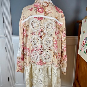 May include: A long-sleeved, beige floral dress with a cream-colored lace back panel and hem. The dress features a button-down front and a collared neckline. The floral pattern includes pink and red roses.