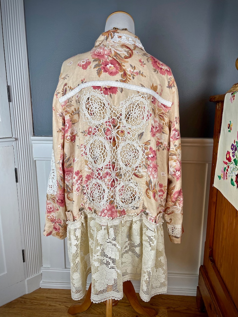 May include: A long-sleeved, beige floral dress with a cream-colored lace back panel and hem. The dress features a button-down front and a collared neckline. The floral pattern includes pink and red roses.
