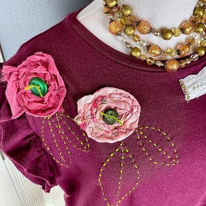 May include: A maroon t-shirt with a ruffled neckline and two pink fabric flowers with green buttons. The shirt has yellow stitching in the shape of leaves around the flowers.