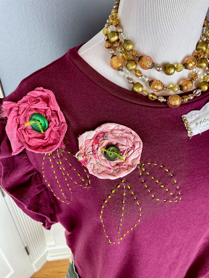 May include: A maroon t-shirt with a ruffled neckline and two pink fabric flowers with green buttons. The shirt has yellow stitching in the shape of leaves around the flowers.