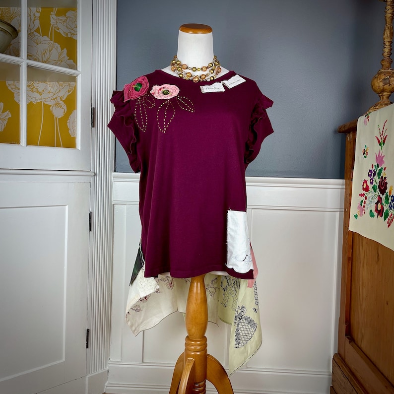 May include: A burgundy t-shirt with a floral applique and a white lace trim. The shirt is on a mannequin and has a high-low hemline.