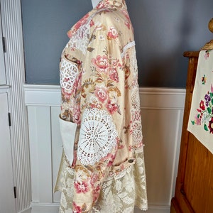May include: A floral print coat with pink and red flowers on a cream background. The coat features white lace and crochet details on the sleeves, pockets, and hem. The coat is displayed on a mannequin.