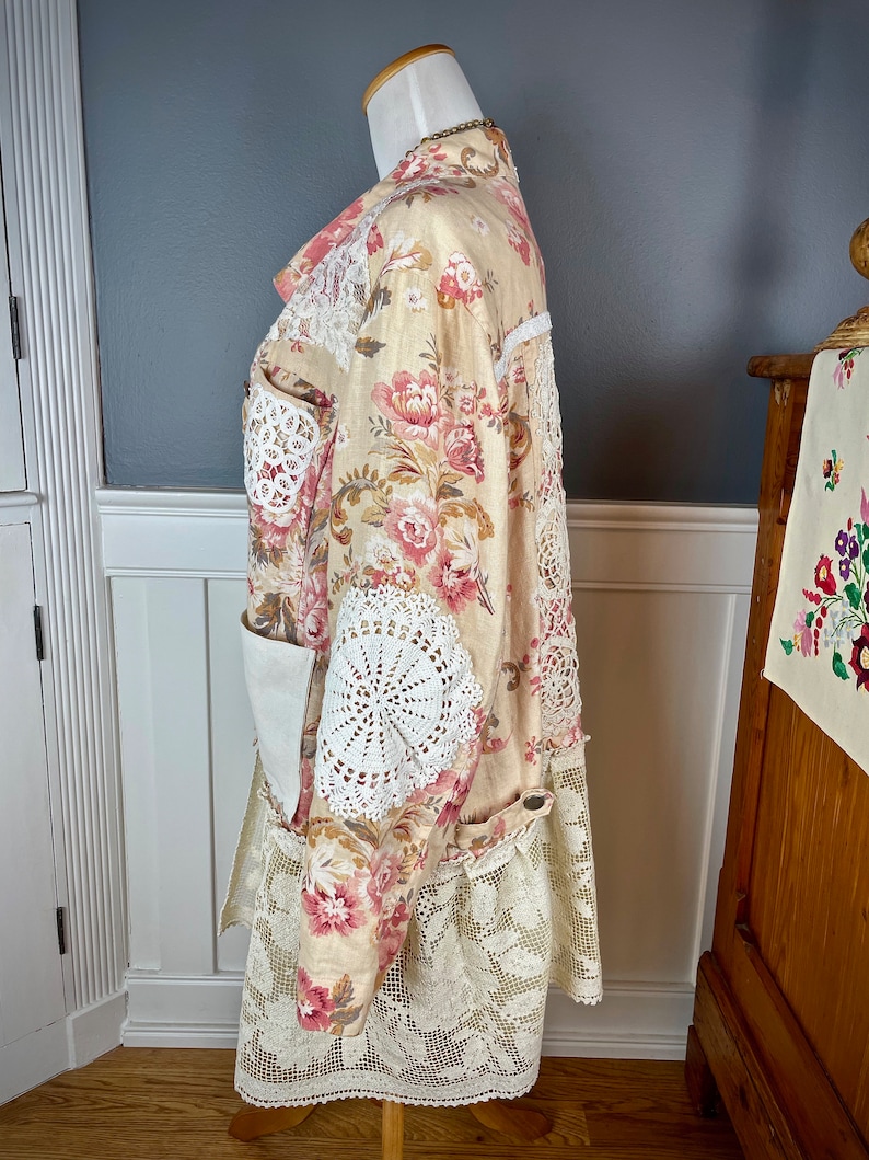 May include: A floral print coat with pink and red flowers on a cream background. The coat features white lace and crochet details on the sleeves, pockets, and hem. The coat is displayed on a mannequin.