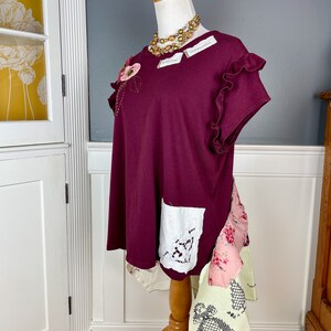 May include: A maroon t-shirt with a floral embellishment on the left shoulder, a white lace pocket on the right side, and a layered patchwork skirt. The shirt has ruffled sleeves and a relaxed fit.
