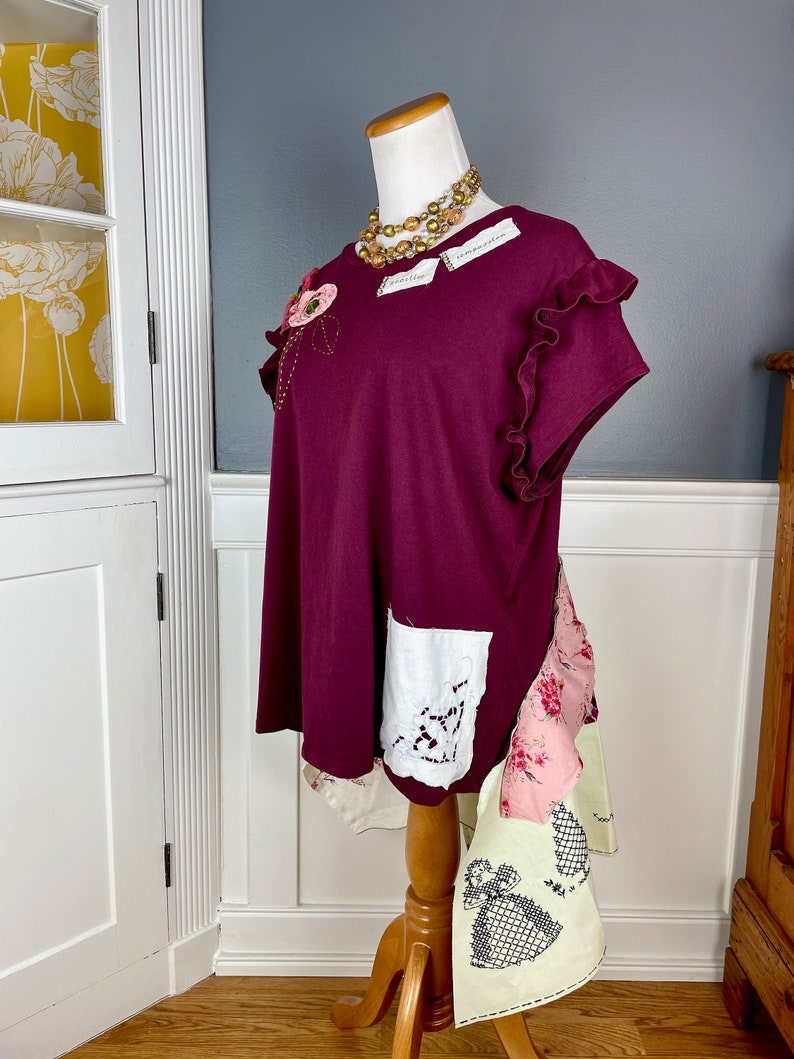 May include: A maroon t-shirt with a floral embellishment on the left shoulder, a white lace pocket on the right side, and a layered patchwork skirt. The shirt has ruffled sleeves and a relaxed fit.