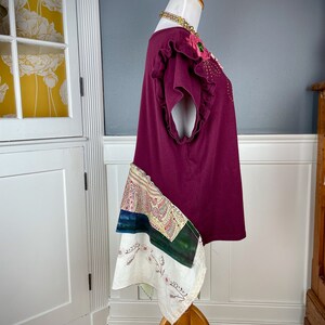 May include: A maroon t-shirt with a floral embellishment on the neckline and a patchwork skirt. The skirt is made of different fabrics in shades of white, green, and blue.