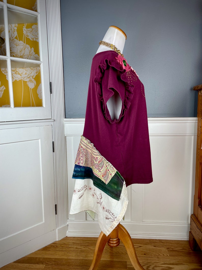 May include: A maroon t-shirt with a floral embellishment on the neckline and a patchwork skirt. The skirt is made of different fabrics in shades of white, green, and blue.