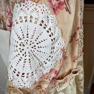 May include: A floral patterned garment with a cream background and pink flowers. White crocheted doilies are sewn onto the sleeves. The garment has a pocket and lace trim. A button closure is visible.