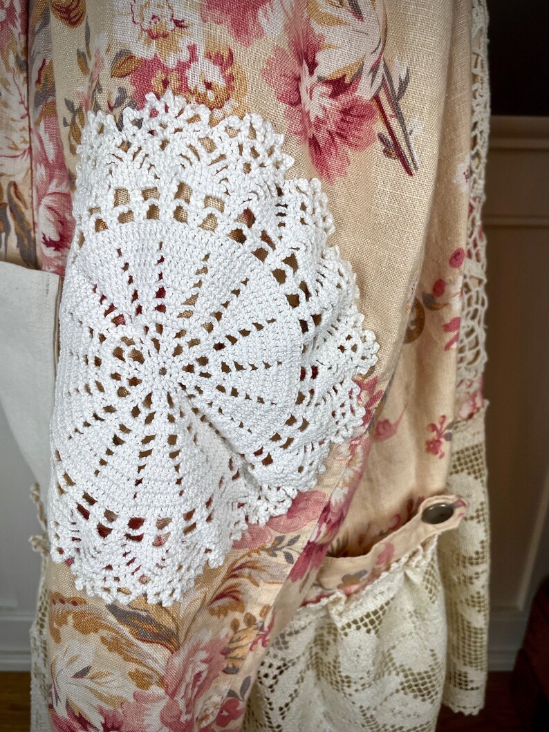 May include: A floral patterned garment with a cream background and pink flowers. White crocheted doilies are sewn onto the sleeves. The garment has a pocket and lace trim. A button closure is visible.
