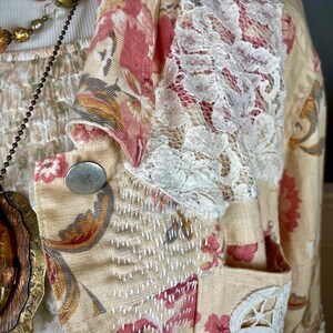 May include: A cream-colored blazer with a floral pattern in pink and red. The blazer features white lace accents on the sleeves and pocket, with silver buttons and decorative stitching. A necklace with gold and clear beads is visible.
