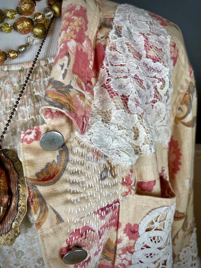 May include: A cream-colored blazer with a floral pattern in pink and red. The blazer features white lace accents on the sleeves and pocket, with silver buttons and decorative stitching. A necklace with gold and clear beads is visible.