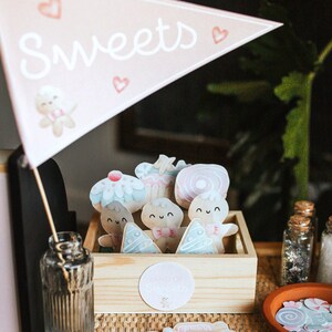 May include: A wooden box with a pink and white sign that says "Sweets" and a variety of colourful, cartoon-style cookies and cupcakes inside. The box is sitting on a table with a brown wicker surface. There are also several small, colourful stickers on the table.