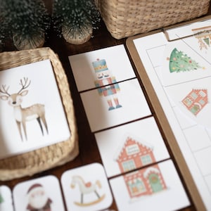 May include: A collection of Christmas-themed educational materials including a woven basket filled with illustrated cards of Santa and reindeer, small bottle brush trees, a wooden house cutout, and a clipboard with a game spinner.