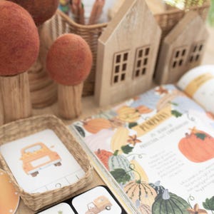 May include: A collection of autumn-themed items including felted wool trees with brown trunks, two wooden house structures, a small woven basket containing a card with a truck illustration, and an open book featuring pumpkin illustrations.