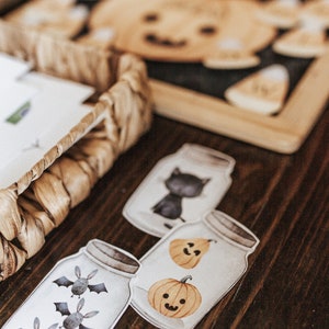 May include: Halloween-themed jar-shaped cutouts with illustrations of bats, pumpkins, and a black cat. The cutouts are arranged on a wooden surface, with a woven basket and pumpkin-shaped cutouts in the background.