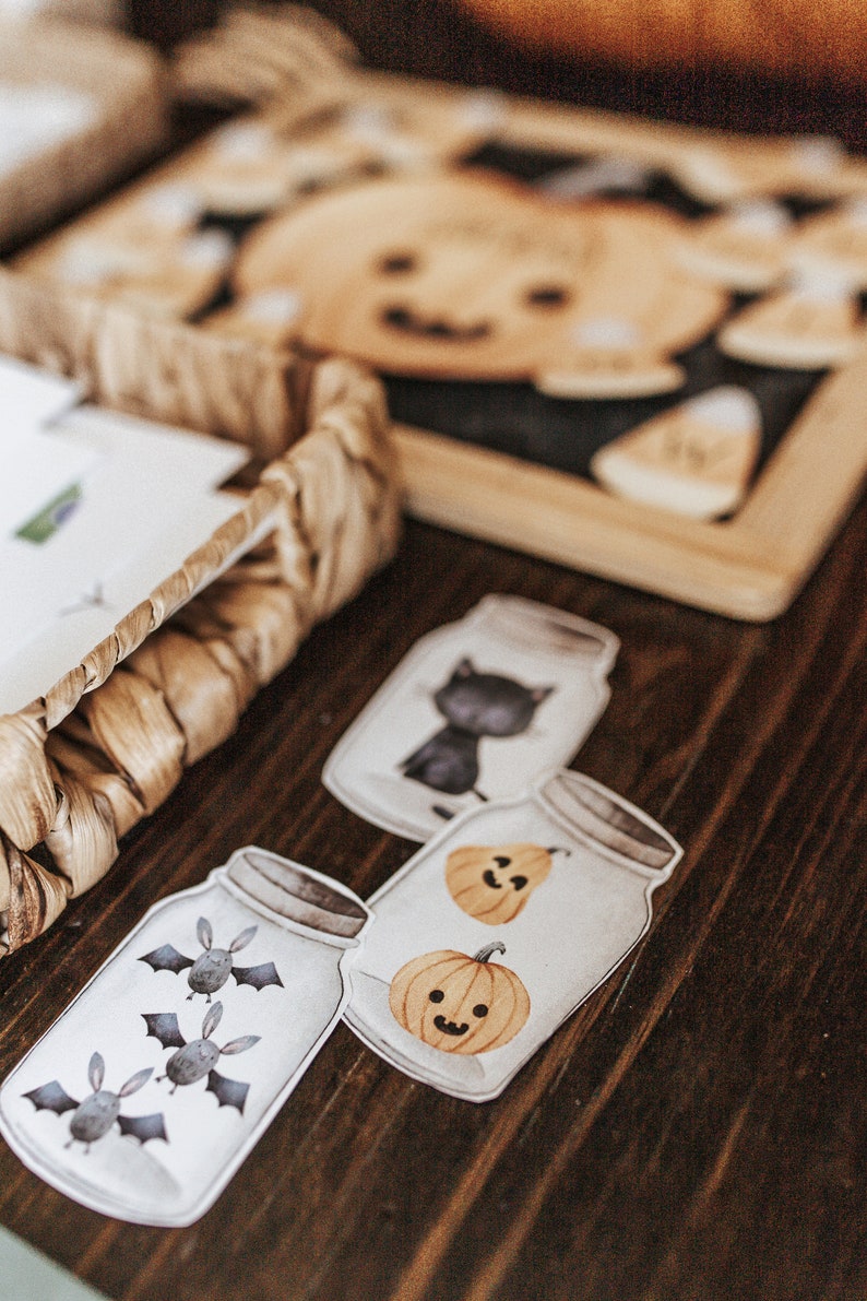 May include: Halloween-themed jar-shaped cutouts with illustrations of bats, pumpkins, and a black cat. The cutouts are arranged on a wooden surface, with a woven basket and pumpkin-shaped cutouts in the background.