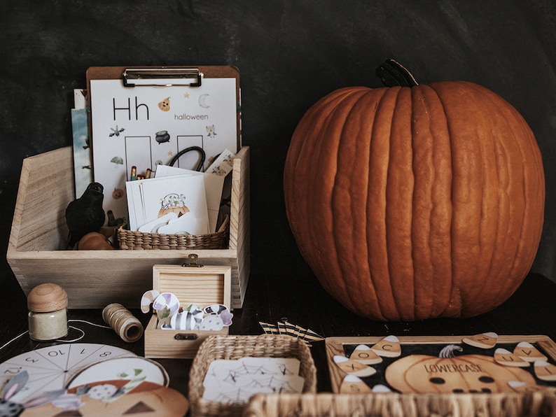 May include: A large orange pumpkin next to a wooden bin filled with Halloween-themed educational materials. The bin contains flashcards, a clipboard with a worksheet, and decorative items. A small wooden box and a basket are also visible.