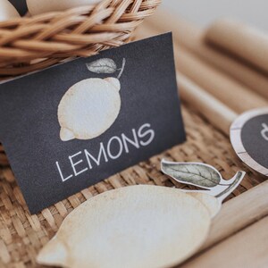 May include: A black card with the word "LEMONS" in white, and a watercolour illustration of a lemon. A wicker basket with eggs is in the background. A wooden dowel is in the foreground. A cut-out lemon is also visible.