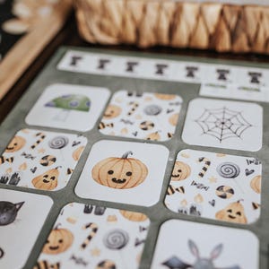 May include: Close-up of Halloween-themed cards with illustrations of a black cat, pumpkin, spider web, and mushroom. The cards have a white background with orange, black, and green accents. A woven basket is in the background.