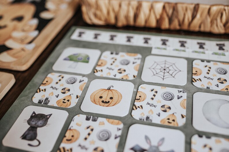 May include: Close-up of Halloween-themed cards with illustrations of a black cat, pumpkin, spider web, and mushroom. The cards have a white background with orange, black, and green accents. A woven basket is in the background.