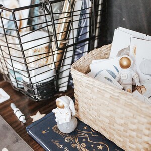 May include: A white astronaut figurine with a gold helmet sits in a woven basket filled with cards featuring space-themed illustrations. A black wire basket filled with craft supplies sits next to the woven basket. A blue book with gold stars and the text "Andy Warhol" is visible in the foreground.
