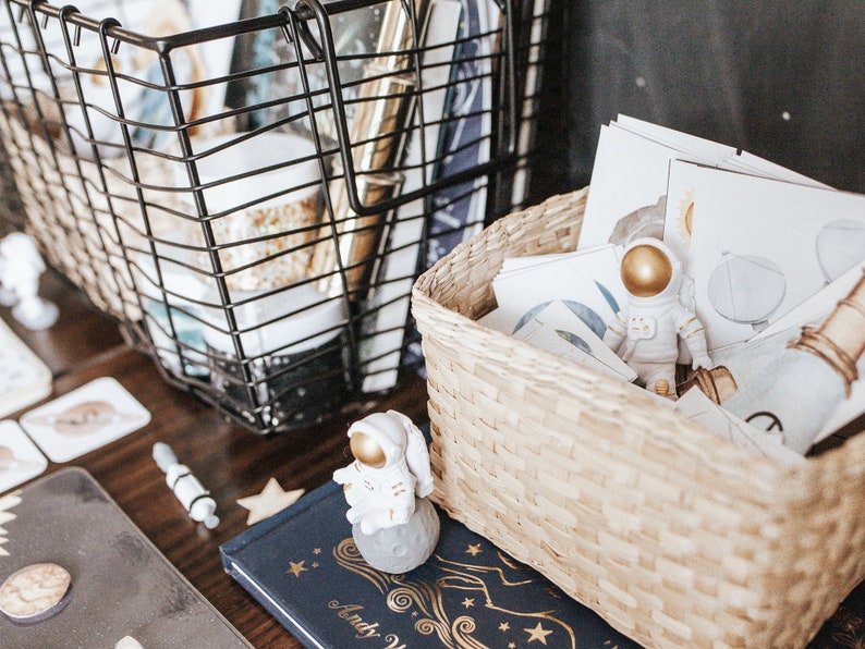 May include: A white astronaut figurine with a gold helmet sits in a woven basket filled with cards featuring space-themed illustrations. A black wire basket filled with craft supplies sits next to the woven basket. A blue book with gold stars and the text "Andy Warhol" is visible in the foreground.