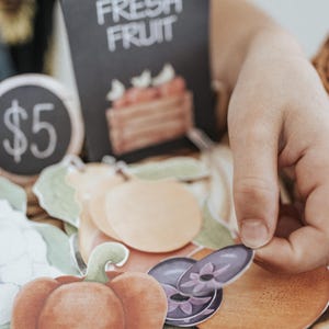May include: A selection of paper fruit and vegetable cutouts, including a red pepper, orange slices, and purple aubergine. A sign reads "FRESH FRUIT" and another shows the price of &pound;5.00.
