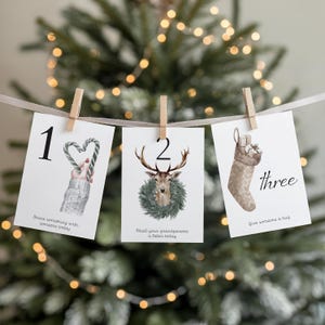 May include: Three white cards, numbered 1, 2, and three, suspended from a string with wooden pegs. Each card displays a watercolour illustration: a candy cane heart, a deer with a wreath, and a Christmas stocking. Text on the cards includes phrases like "Share something" and "Give someone a hug."