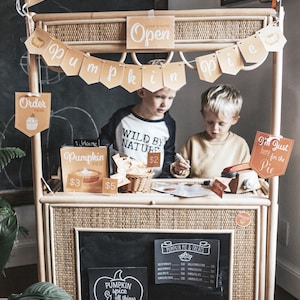 May include: Two children are playing at a pretend pumpkin pie stand. The stand is made of wicker and has a chalkboard sign that says "Pumpkin Pie & Treats". There are also signs that say "Pumpkin Spice & all things nice", "Wild by Nature", and "I'm just here for the pie".