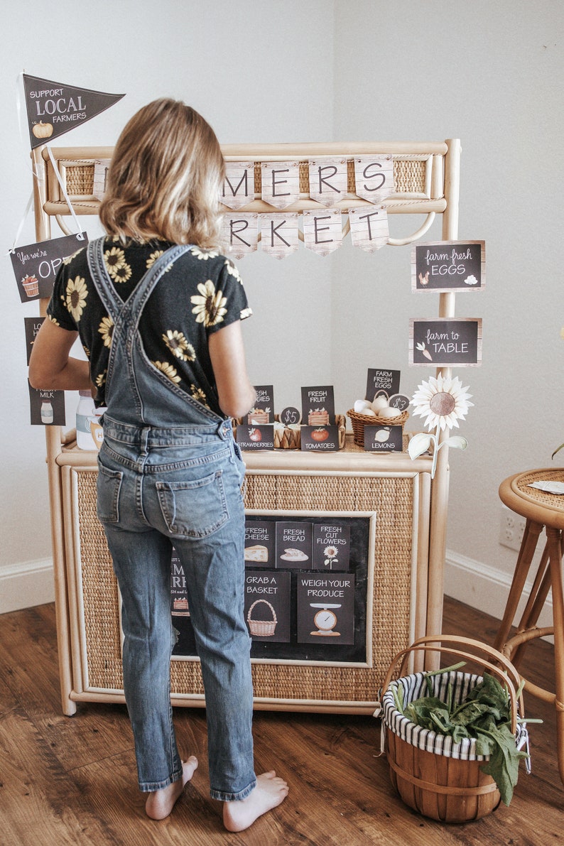 May include: A child stands in front of a farmers market stall made of woven wicker. The stall has signs for fresh produce, eggs, and flowers. The banner reads "FARMERS MARKET". A basket of greens sits in front.