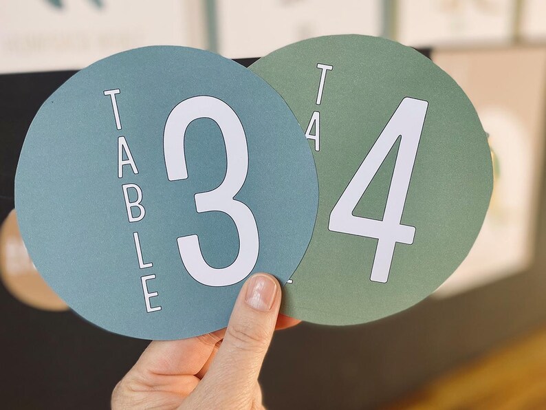 May include: Two round teal and green table number cards with white numbers. The teal card says "TABLE" and has the number "3" on it. The green card has the number "4" on it.