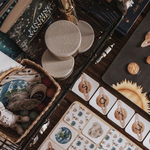May include: A black wire basket filled with a variety of items, including a book titled "Finding Wonders", a set of small white jars with the word "Free" written on them, and a set of colorful wooden circles with numbers on them. The basket is sitting on a wooden table with a black and white solar system chart and a set of alphabet cards.