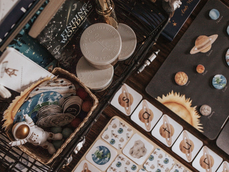 May include: A black wire basket filled with a variety of items, including a book titled "Finding Wonders", a set of small white jars with the word "Free" written on them, and a set of colorful wooden circles with numbers on them. The basket is sitting on a wooden table with a black and white solar system chart and a set of alphabet cards.