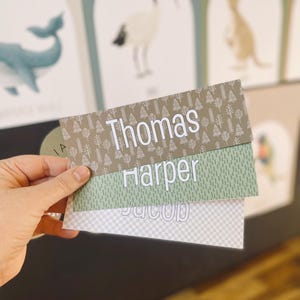 Könnte beinhalten: Drei personalisierte Namensschilder mit Waldmotiv. Das oberste Schild hat einen braunen Hintergrund mit weißen Bäumen und dem Namen "Thomas". Das mittlere Schild hat einen grünen Hintergrund mit weißen Linien und dem Namen "Harper". Das unterste Schild hat einen weißen Hintergrund mit einem grauen Karomuster und dem Namen "Jacob".