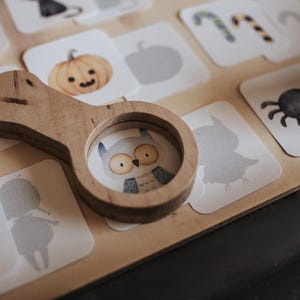May include: A wooden magnifying glass rests on a collection of Halloween-themed cards. The cards feature illustrations of a pumpkin, owl, spider, and other spooky images. The magnifying glass has a circular lens with an owl illustration.