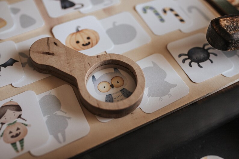 May include: A wooden magnifying glass rests on a collection of Halloween-themed cards. The cards feature illustrations of a pumpkin, owl, spider, and other spooky images. The magnifying glass has a circular lens with an owl illustration.