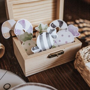 May include: A wooden box with a hinged lid, filled with colorful paper candy cutouts. The candies feature various designs, including stripes, polka dots, and swirls in shades of purple, green, and brown. The box is made of light-colored wood.