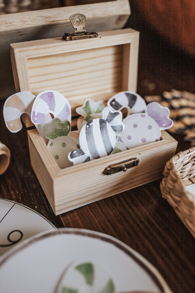 May include: A wooden box with a hinged lid, filled with colorful paper candy cutouts. The candies feature various designs, including stripes, polka dots, and swirls in shades of purple, green, and brown. The box is made of light-colored wood.