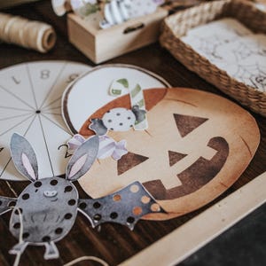 May include: A collection of Halloween-themed paper cutouts and decorations. Includes a pumpkin, bat, and candy corn, along with a circular game piece and a small woven basket. The color palette features warm tones and watercolor-style illustrations.