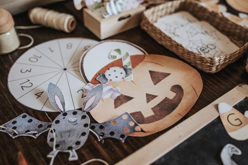 May include: A collection of Halloween-themed paper cutouts and decorations. Includes a pumpkin, bat, and candy corn, along with a circular game piece and a small woven basket. The color palette features warm tones and watercolor-style illustrations.