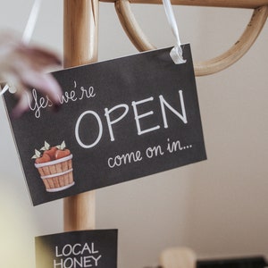 May include: A black chalkboard sign with the words "Yes we're OPEN come on in..." is suspended from a wooden display. The sign has a watercolour illustration of a basket of apples. A smaller sign below reads "LOCAL HONEY".