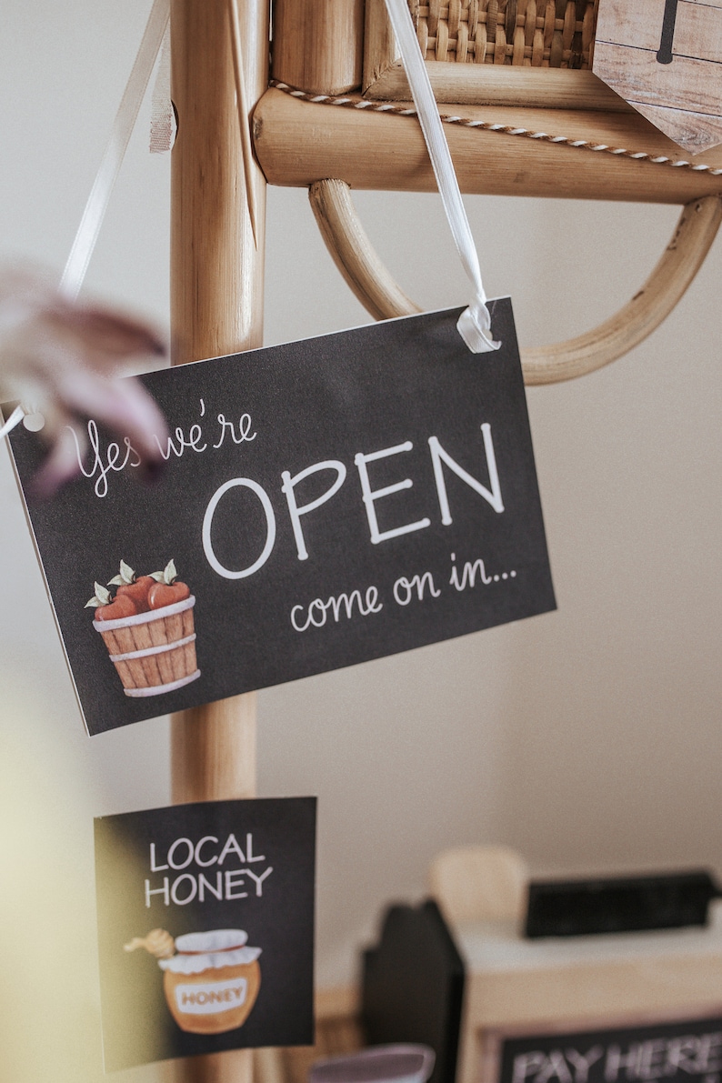 May include: A black chalkboard sign with the words "Yes we're OPEN come on in..." is suspended from a wooden display. The sign has a watercolour illustration of a basket of apples. A smaller sign below reads "LOCAL HONEY".