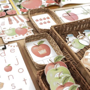 May include: A collection of educational materials featuring apple-themed illustrations, including printed cards, a puzzle, and alphabet sheets. Several woven baskets contain die-cut apple shapes in red and green, alongside small cards with apple graphics.