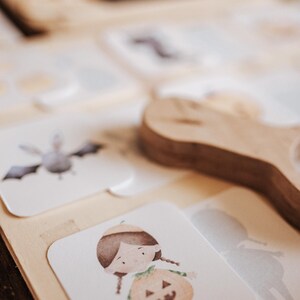 May include: Wooden Halloween-themed puzzle with illustrated cards. The cards feature a bat, a girl in a pumpkin costume, and other Halloween-related images. A wooden handle is visible in the background. The puzzle is designed for children.