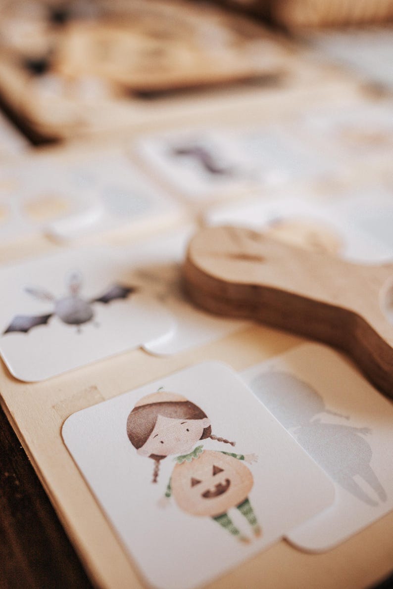 May include: Wooden Halloween-themed puzzle with illustrated cards. The cards feature a bat, a girl in a pumpkin costume, and other Halloween-related images. A wooden handle is visible in the background. The puzzle is designed for children.
