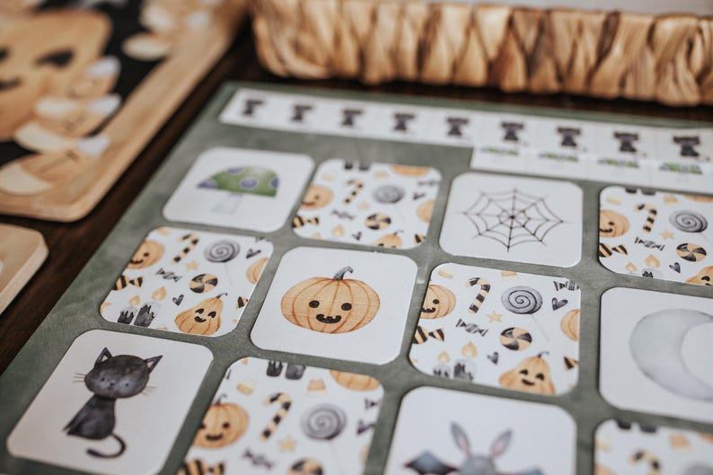 May include: Close-up of Halloween-themed cards with illustrations of a black cat, pumpkin, spider web, and mushroom. The cards have a white background with orange, black, and green accents. A wooden tray and woven basket are in the background.