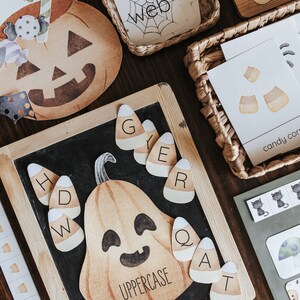 May include: Halloween-themed educational materials. Includes a pumpkin-shaped chalkboard with candy corn letter cards, a pumpkin cutout, and cards with the word "web" and candy corn illustrations. The materials are arranged on a wooden surface.