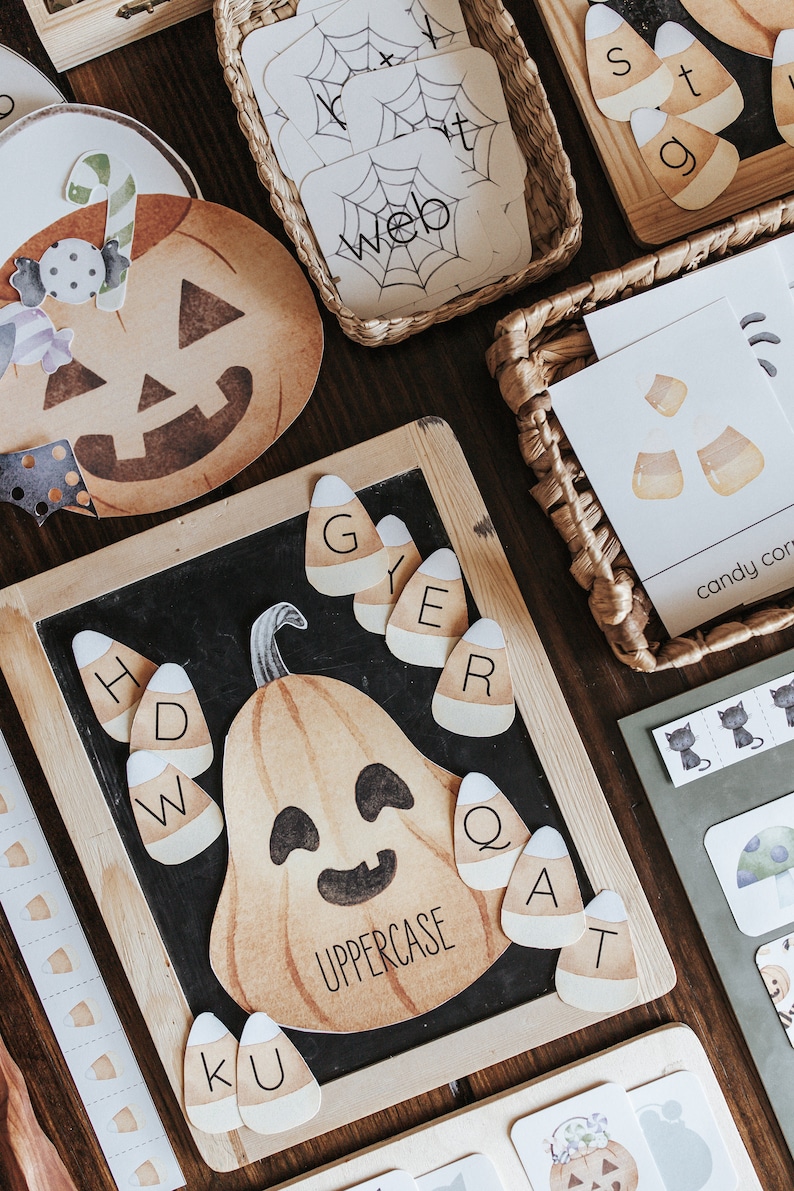 May include: Halloween-themed educational materials. Includes a pumpkin-shaped chalkboard with candy corn letter cards, a pumpkin cutout, and cards with the word "web" and candy corn illustrations. The materials are arranged on a wooden surface.