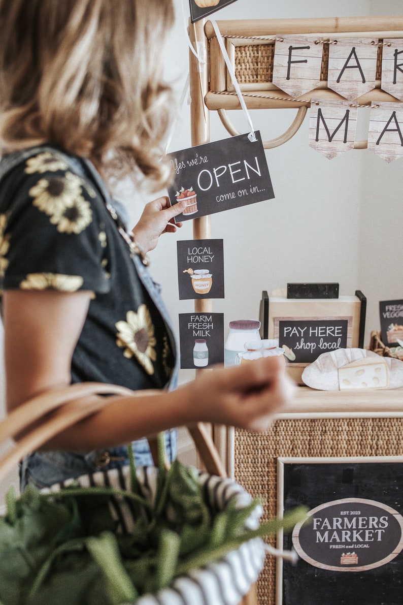 May include: A farmers market scene with signs that read "OPEN", "Local Honey", and "Farm Fresh Milk". A person holds a basket of produce. Banners spell out "FARM MARKET". A sign says "PAY HERE shop local".